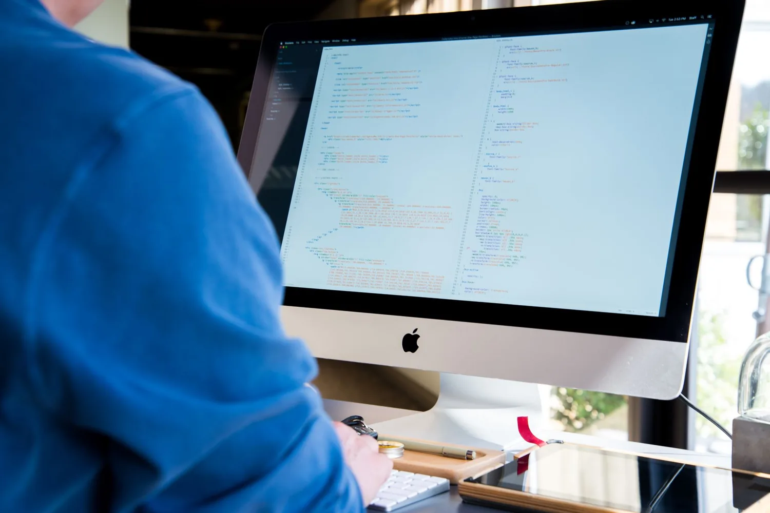 Pessoa de moletom azul digitando em teclado diante de monitor com código-fonte colorido
