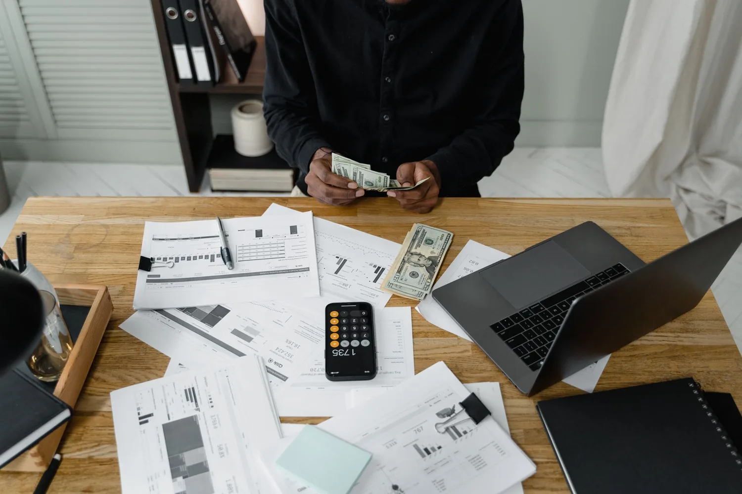 Mãos contando dinheiro sobre uma mesa de madeira com relatórios financeiros, notebook e celular com calculadora aberta